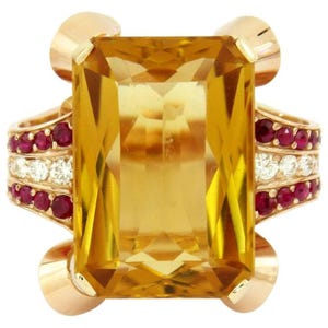 May include: A gold ring featuring a large, rectangular, yellow gemstone as the centerpiece. The ring is accented with rows of small, red gemstones and white diamonds. The design includes gold accents on the sides, creating a luxurious look.