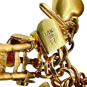 May include: Close-up of a gold-colored charm bracelet with a key and heart-shaped charms. The bracelet has a clasp with the inscription "14K" and a crown symbol. The links are a series of interlocking circles.