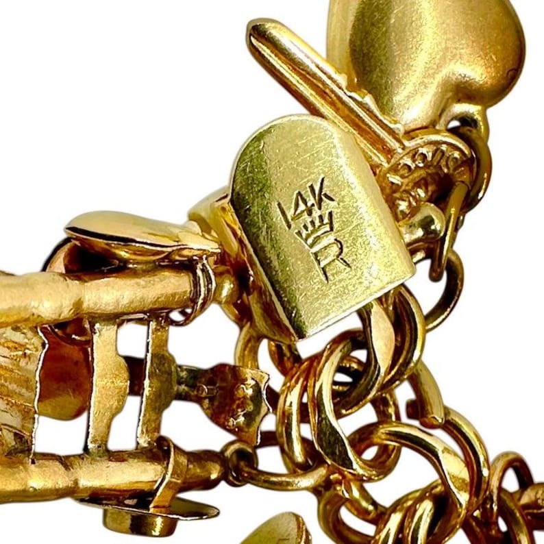 May include: Close-up of a gold-colored charm bracelet with a key and heart-shaped charms. The bracelet has a clasp with the inscription "14K" and a crown symbol. The links are a series of interlocking circles.