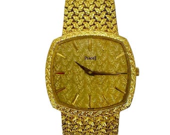 May include: A gold-toned Piaget wristwatch with a textured dial and woven band. The watch face is square with black hands and the Piaget logo. The band is wide and flexible, with a matching textured pattern. The watch is a luxury item.