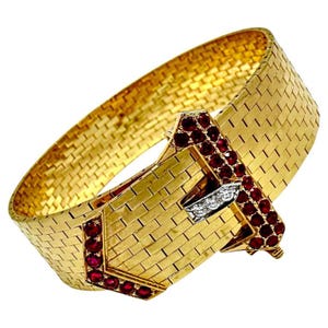 May include: A wide, gold-toned bracelet with a woven brickwork design. The clasp features a geometric design with red gemstones and a small section of white gemstones. The bracelet is a statement piece of jewelry.
