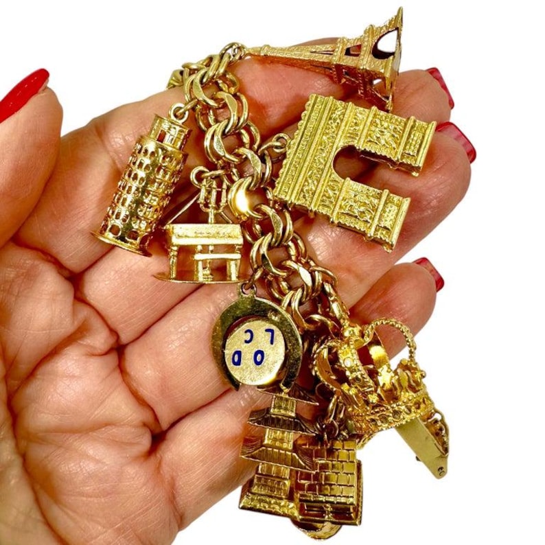 May include: A gold charm bracelet featuring miniature replicas of famous landmarks. The charms include the Eiffel Tower, the Leaning Tower of Pisa, and a London-themed charm. The bracelet is held in a hand against a white background.