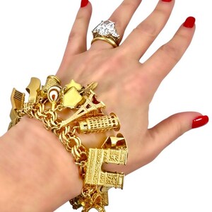 May include: A gold charm bracelet with various travel-themed charms, including the Eiffel Tower and a leaning tower. A large diamond ring is also visible. The person's nails are painted red.