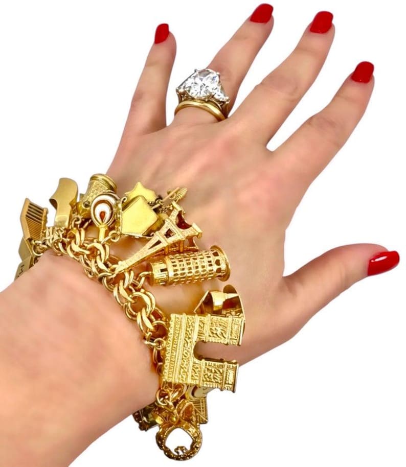 May include: A gold charm bracelet with various travel-themed charms, including the Eiffel Tower and a leaning tower. A large diamond ring is also visible. The person's nails are painted red.