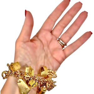 May include: A gold charm bracelet with various charms, including a piano, a measuring tape, a chair, and a teapot. The bracelet is made of gold-colored metal and is displayed on a person's wrist.