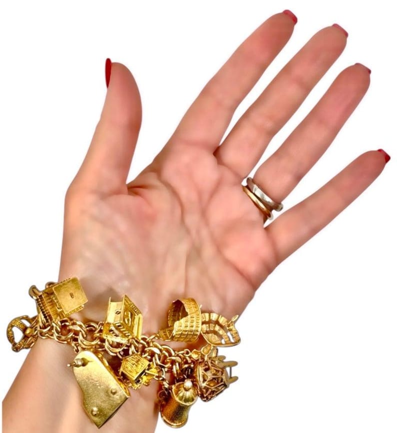 May include: A gold charm bracelet with various charms, including a piano, a measuring tape, a chair, and a teapot. The bracelet is made of gold-colored metal and is displayed on a person's wrist.