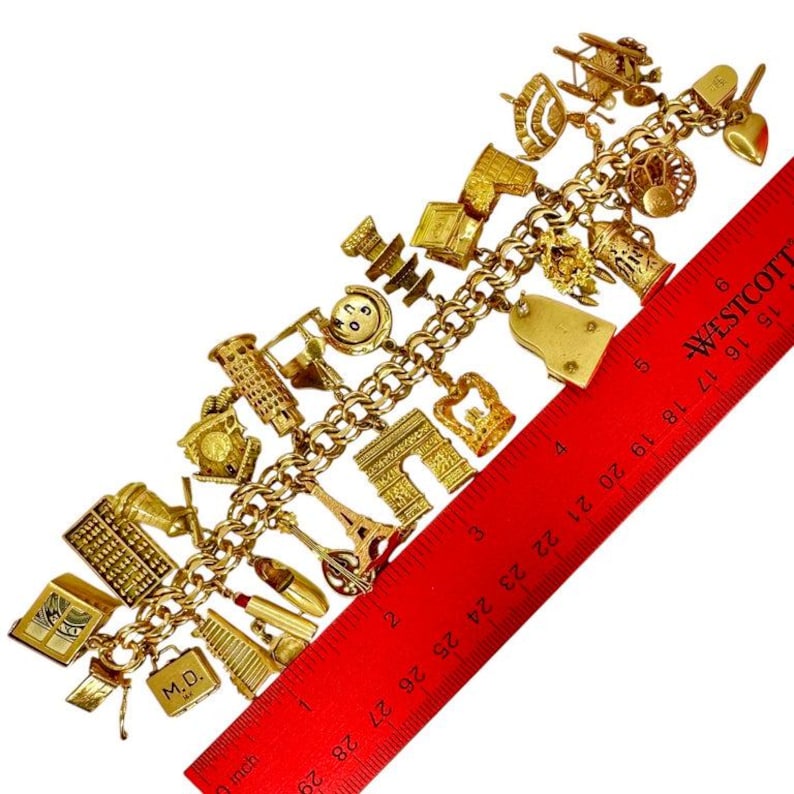 May include: Gold-toned charm bracelet featuring various miniature charms, including the Eiffel Tower, a crown, and a piano. The bracelet is displayed with a red ruler, showing measurements in inches.