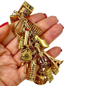 May include: A gold-toned charm bracelet featuring miniature travel-themed charms. Charms include a miniature Eiffel Tower, a leaning tower of Pisa, a Dutch clog, a cuckoo clock, and a miniature suitcase with the initials "M.D."