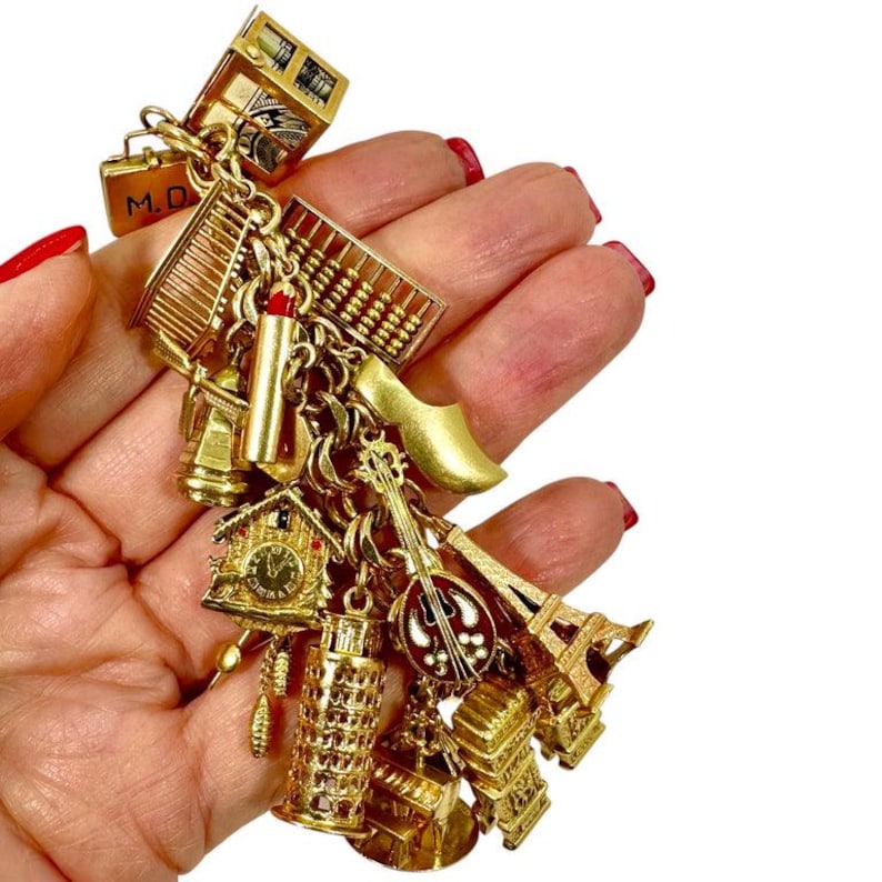May include: A gold-toned charm bracelet featuring miniature travel-themed charms. Charms include a miniature Eiffel Tower, a leaning tower of Pisa, a Dutch clog, a cuckoo clock, and a miniature suitcase with the initials "M.D."