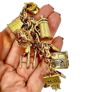 May include: A gold charm bracelet with various charms, including a beer stein, a miniature house, a suitcase with "M.D." engraved, and a small ladder. The charms are intricately detailed and connected by a gold chain.