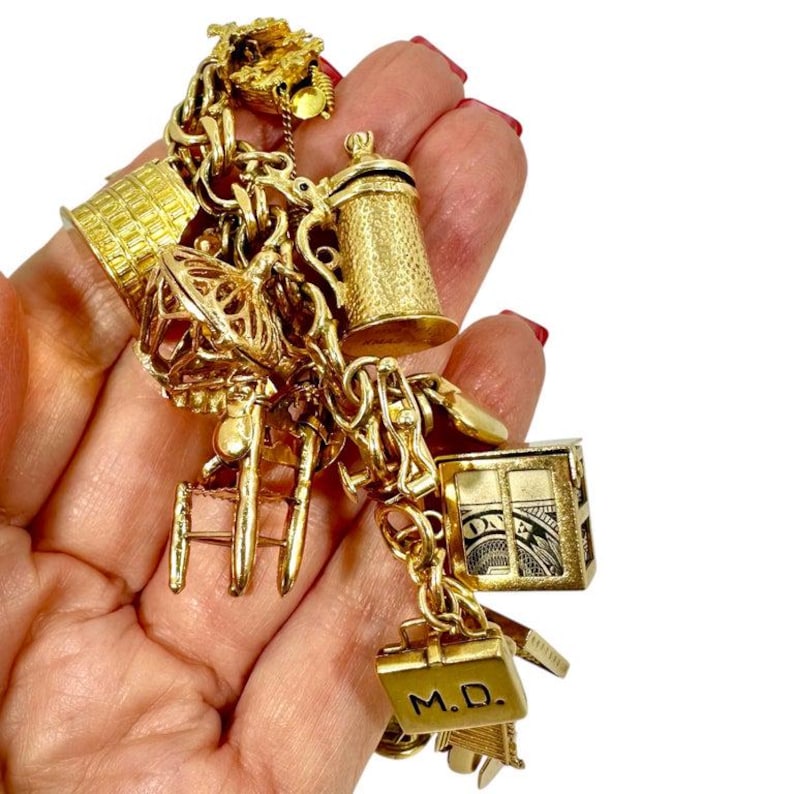 May include: A gold charm bracelet with various charms, including a beer stein, a miniature house, a suitcase with "M.D." engraved, and a small ladder. The charms are intricately detailed and connected by a gold chain.