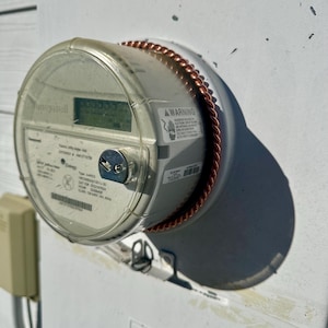 May include: A Honeywell electric meter with a clear plastic cover is mounted on a white wall. The meter has a digital display and a copper-colored wire around the edge. A warning label is visible on the meter.