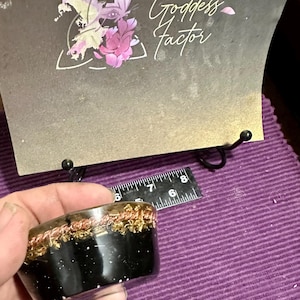 May include: A black and gold orgonite energy device with copper and gold accents. The device is held in a hand, with a ruler and a card with the text "Goddess Factor" in the background. The card has a floral and butterfly design.