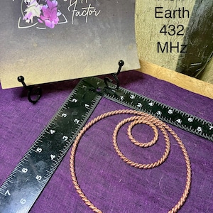 May include: Two copper wire rings with a twisted texture, one large and one small, placed next to a black L-shaped ruler showing measurements in inches. A sign behind them reads Goddess Factor New Earth 432 MHz.