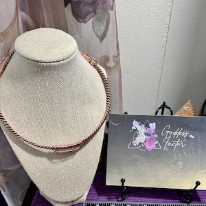 May include: A copper choker necklace with a twisted rope design, displayed on a mannequin head. The necklace features a thicker central section. A business card with "Goddess Factor" is in the foreground.