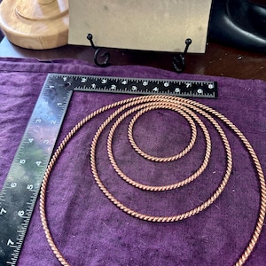 May include: Four copper-colored, twisted metal rings of varying sizes are arranged on a purple cloth. A ruler is placed next to the rings for scale. A sign with the text "Goddess factor" is in the background.