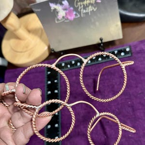May include: Copper-colored wire eyeglasses with a twisted rope design. The glasses have four circular frames connected by thin metal. A ruler and a card with the text "Goddess Factor" are in the background.