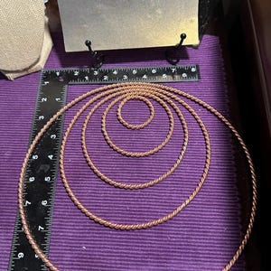 Puede incluir: Aros de metal color cobre de varios tamaños con diseño de cuerda trenzada sobre una superficie morada. Una regla se encuentra junto a los aros. Un letrero con el texto "Goddess Factor" se encuentra en el fondo.