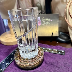 Puede incluir: Un vaso transparente con un diseño en espiral, lleno de agua, se encuentra sobre un posavasos redondo con alambre de cobre y escamas doradas. También se ven una regla y un pequeño letrero con texto.