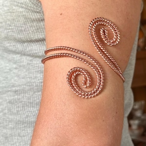 May include: A copper-colored arm cuff featuring a double-strand twisted wire design. The metal wire forms two opposing spiral ends that curve outward, creating an open-ended, adjustable band that wraps around the upper arm with a metallic finish.