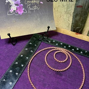 May include: Two copper-colored twisted wire rings of different sizes are displayed next to an L-shaped ruler. A card behind them features the text Cosmic Connector 528 MHz and Goddess Factor. The ruler shows the larger ring is 6 inches wide.