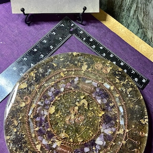 May include: A circular resin disc featuring embedded purple amethyst stones, copper wire coils, and gold leaf flakes. A black L-shaped ruler is placed across the top, showing a diameter of approximately 8 inches. A sign above reads Goddess Factor.