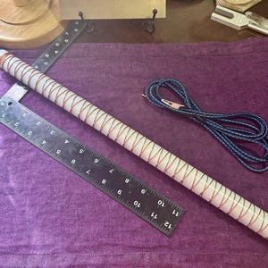 May include: A white, cylindrical object wrapped with copper wire, approximately 12 inches long, lies on a purple surface. A black ruler, a blue and black cable, and other objects are also present.