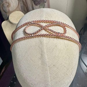 May include: A copper-colored metal headband featuring a twisted rope texture. The design includes a central infinity symbol shape formed by the continuous metal wire. The headband is displayed on a beige fabric-covered mannequin head form.