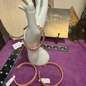 May include: Copper-colored braided bracelets, some displayed on a white hand-shaped jewelry holder. The bracelets are on a purple surface, with a ruler and a card that says "Goddess Factor".