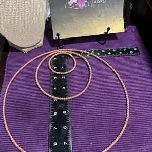 May include: Three copper-colored, circular jewelry pieces of varying sizes are displayed on a purple surface next to a ruler. A sign with the text "Goddess Factor" is in the background.