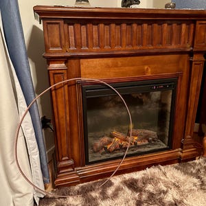 May include: A dark wood fireplace with a black glass front. The fireplace has a faux fire with glowing logs. A copper-colored metal hoop is in the foreground. The fireplace is set against a wall with a curtain.