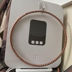 May include: A white electronic device with a black screen and control buttons, featuring a copper-colored, twisted metal ring. The device has the T-Mobile logo and is mounted on a wall.