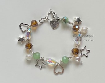 Puede incluir: Una pulsera de cuentas plateada con cierre de palanca. La pulsera presenta cuentas marrones, verdes e iridiscentes, junto con dijes de estrella y corazón. Un pequeño dije de corazón dice "Made with Love".