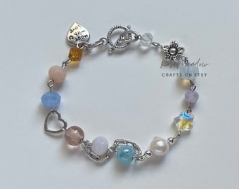 Puede incluir: Una pulsera de cuentas con una variedad de cuentas coloridas, incluyendo rosa, azul y amarillo. La pulsera tiene un cierre plateado y un dije en forma de corazón. El texto "Made with Love" está en un dije en forma de corazón. La pulsera está sobre una superficie blanca.