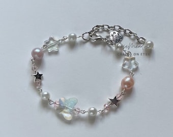 Puede incluir: Un delicado brazalete de cadena plateada con perlas rosas y blancas, estrellas de cristal transparente y un dije de mariposa iridiscente. El brazalete tiene un cierre plateado con un dije de corazón que dice "Rose Meadow Crafts on Etsy".