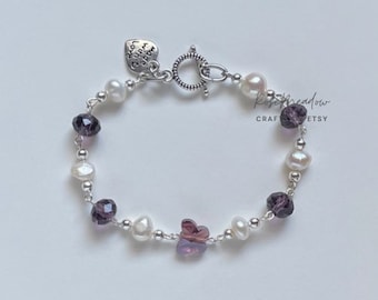 Puede incluir: Un brazalete de plata con perlas blancas, abalorios de mariposa púrpura y un abalorio en forma de corazón que dice "Love you more".