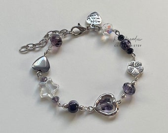May include: A silver charm bracelet with purple and clear beads, silver heart, star, and flower charms. The bracelet has a silver clasp and a tag that reads "Made with Love".