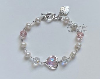 May include: A silver bracelet with white pearls, pink crystals, and a silver heart charm. The charm has a pink iridescent finish. The bracelet has a silver toggle clasp with a heart-shaped tag that reads "and I love you so much".