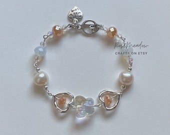 May include: A delicate bracelet featuring a heart-shaped clasp and charms. The bracelet includes iridescent beads, faux pearls, and heart-shaped charms with orange accents. The text "RoseMeadow Crafts on Etsy" is visible.