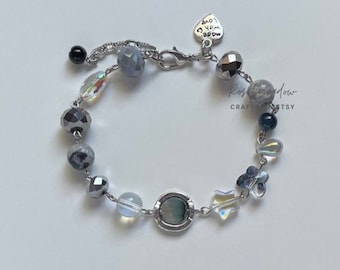 Puede incluir: Un brazalete de plata con una variedad de cuentas, incluyendo cuentas transparentes, grises y negras. El brazalete también incluye un dije de plata con las palabras "Made with Love" en él.