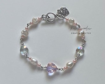 Op de afbeelding: Een delicate armband met een mix van roze en witte parels, iriserende glazen kralen en een hartvormig kristal. De armband heeft een zilveren sluiting en een klein bedeltje met de tekst "Made with Love".