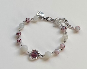 Puede incluir: Un brazalete plateado con cuentas blancas y rojas y un dije de corazón plateado. El dije tiene un centro de vidrio morado. El brazalete tiene un cierre plateado con una etiqueta en forma de corazón que dice "Love Mom".