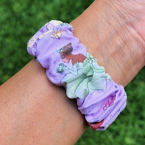 May include: Purple fabric hair scrunchie with a print of Disney princesses, including Tiana, Ariel, and Cinderella.