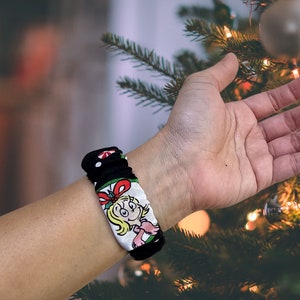 May include: A black and white fabric wristband featuring a cartoon character in a red and green dress. The wristband is worn on a person's wrist.