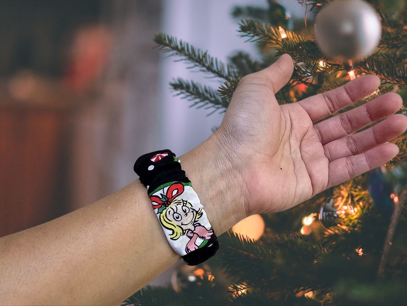 May include: A black and white fabric wristband featuring a cartoon character in a red and green dress. The wristband is worn on a person's wrist.