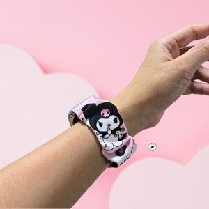Op de afbeelding: Een patroon horlogebandje in zwart, wit en roze, met een cartoon karakter ontwerp. De band is bevestigd aan een zilveren horloge en wordt om de pols gedragen. De achtergrond is lichtroze.