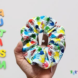 Puede incluir: Un scrunchie blanco con un estampado de huellas de manos de colores. Las huellas de manos son en tonos de rojo, amarillo, azul y verde.