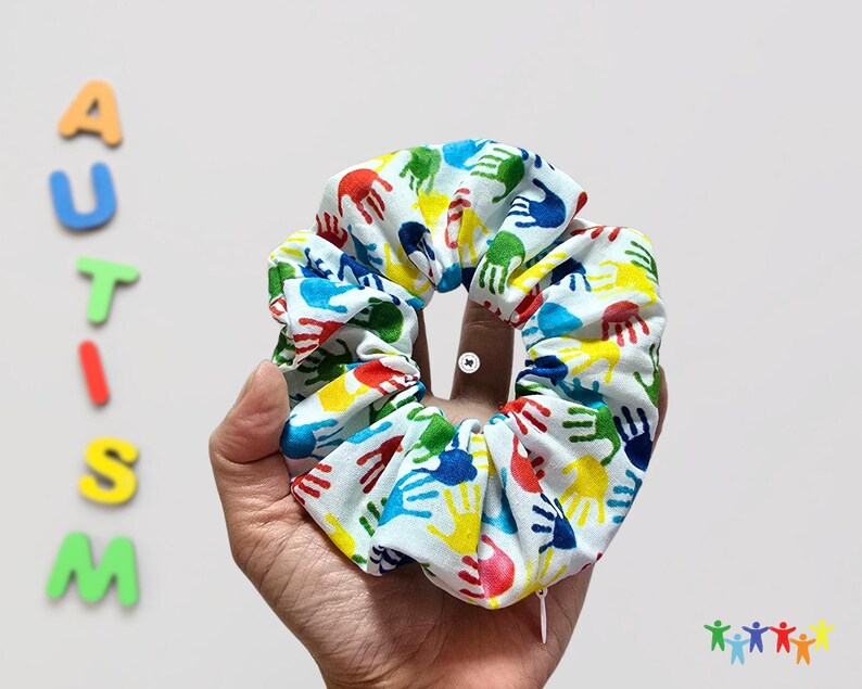 Puede incluir: Un scrunchie blanco con un estampado de huellas de manos de colores. Las huellas de manos son en tonos de rojo, amarillo, azul y verde.