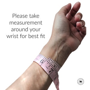 Op de afbeelding: Een roze flexibel meetlint gewikkeld om een pols met de tekst Please take measurement around your wrist for best fit. Het lint toont zwarte markeringen en cijfers, waaronder 6 en 7, die de afmetingen in inches aangeven.