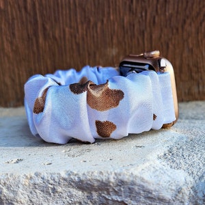 Op de afbeelding: Een horlogeband in scrunchie-stijl met een witte stoffen basis en een bruin koeienvlekkenpatroon. De band is bevestigd aan een ros&eacute;gouden smartwatch-behuizing en heeft een geplooid, elastisch ontwerp dat om de pols past.
